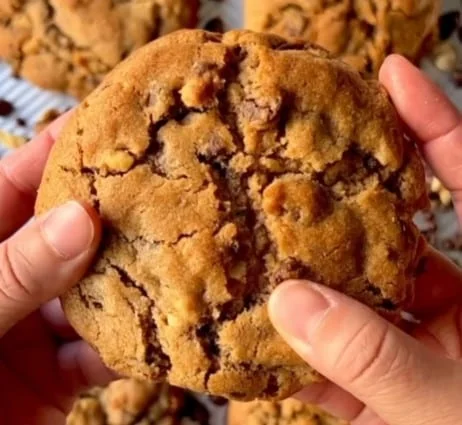 A hand holding a large, cracked Chocolate Chip Walnut Cookie with visible chunks