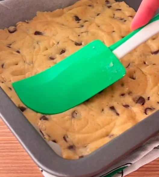 Green spatula spreading cookie dough in a square baking pan