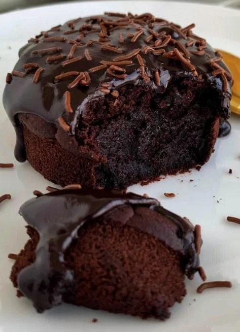 A mini chocolate cake with shiny chocolate glaze and sprinkles, featuring a moist crumb and no layers, with a single piece separated