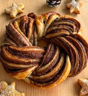 A Nutella pastry wreath sits on a wooden board, accompanied by pine cones and star-shaped cookies, creating a cozy holiday atmosphere