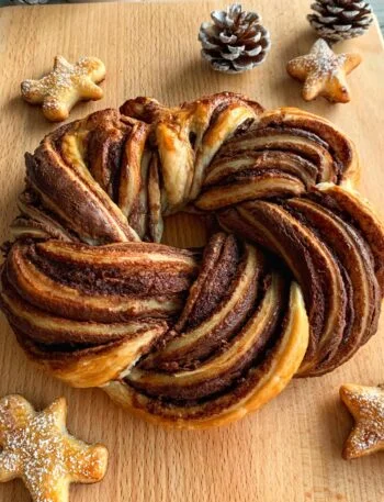 A Nutella pastry wreath sits on a wooden board, accompanied by pine cones and star-shaped cookies, creating a cozy holiday atmosphere