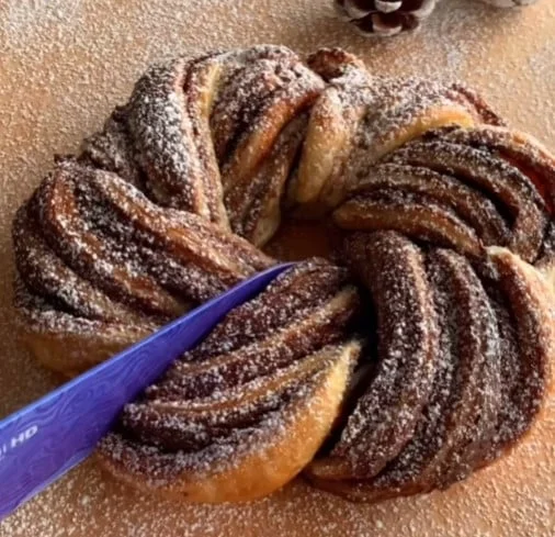 A Nutella pastry wreath, dusted with sugar, with a purple knife cutting into it